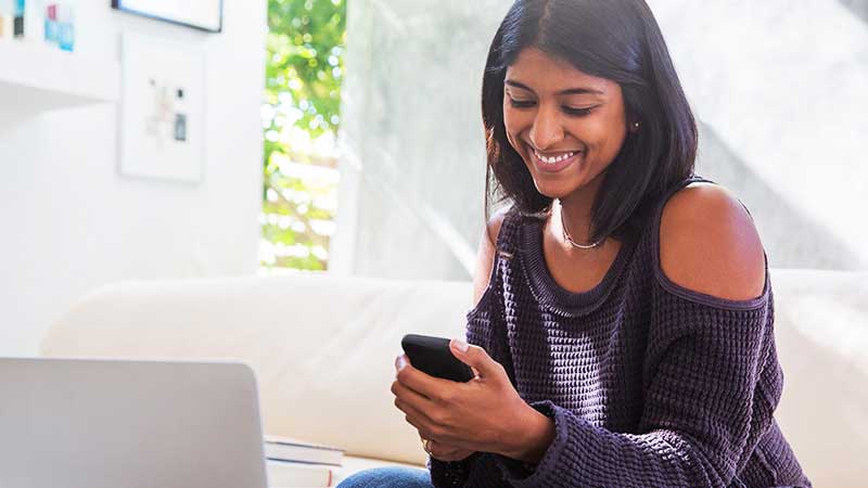 žena-telefonira-800x450 A smiling young woman, sitting on a couch, uses her phone to shop.