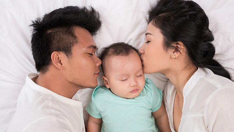 A baby with its eyes closed lying in a bed being kissed by its parents who are lying on both sides.