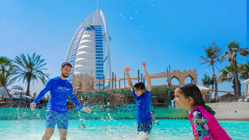 A man and two children play in a water park pool with the Burj Al Arab hotel visible in the background on a sunny day.