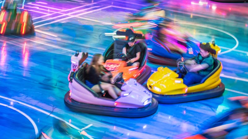 People driving colorful bumper cars indoors with blurred motion, neon lights, and a shiny blue floor, creating a lively and energetic atmosphere.