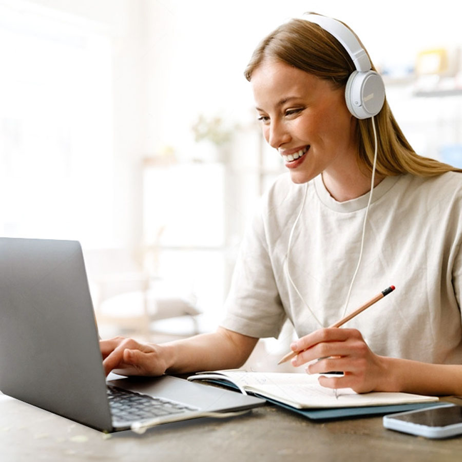 A young woman wearing headphones smiles while using a laptop and taking notes at a bright cafe table.