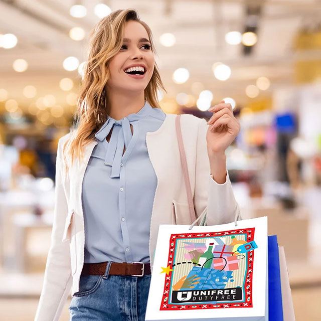 Smiling woman in a mall carrying colorful Duty Free shopping bags, wearing a light blue blouse and white jacket. The scene is bright and cheerful.
