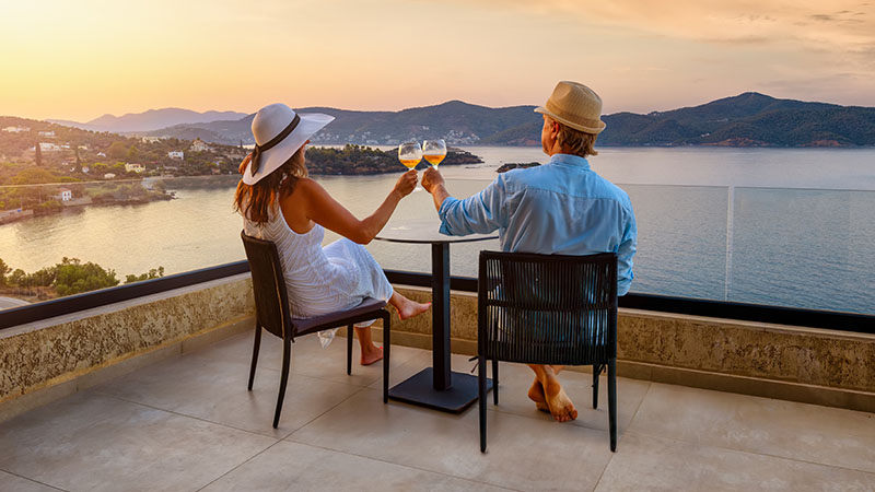 Couple in hats sit on a terrace, clinking wine glasses, overlooking a serene coastal sunset. 