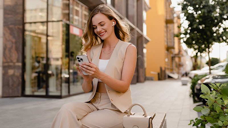 Woman in stylish beige outfit sits on a city bench, smiling while looking at her phone. 