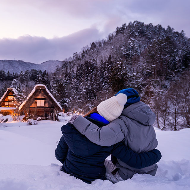 A couple in winter clothing sits on snowy ground, embracing and facing traditional, illuminated cottages against a backdrop of snowy, forested mountains.
