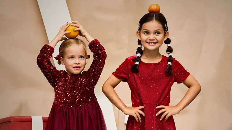 Two girls dressed in red dresses hold an orange on their heads