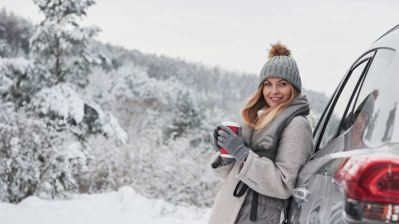 Woman with coffee standing by the car, snowy forests in the background