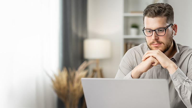 A man wearing glasses and a gray shirt intently looks at his laptop screen in a bright, modern office setting.