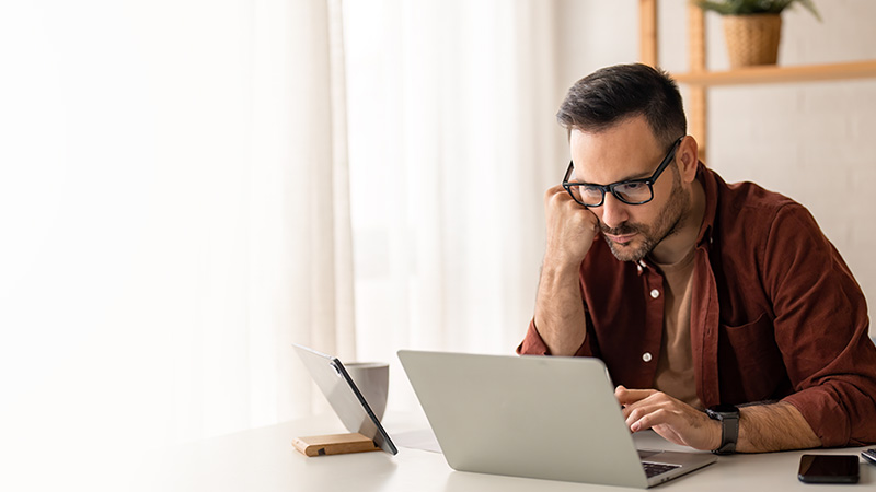 A man with glasses, wearing a red shirt, intently looking at a laptop screen in a bright, modern office setting.